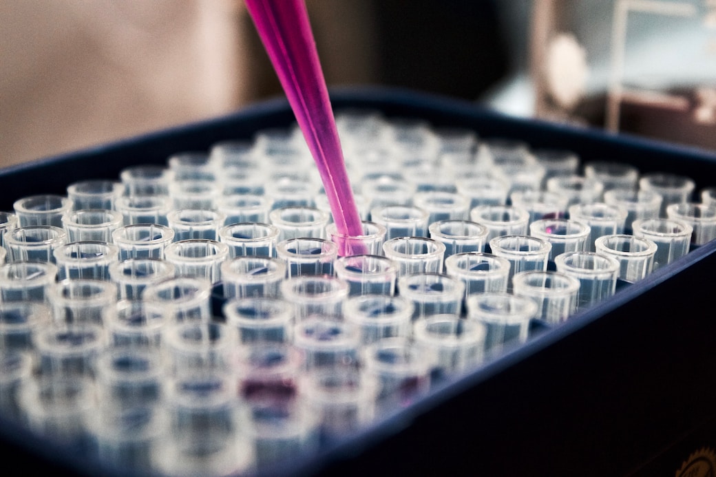 Syringe of purple liquid over a tray of clear test tubes