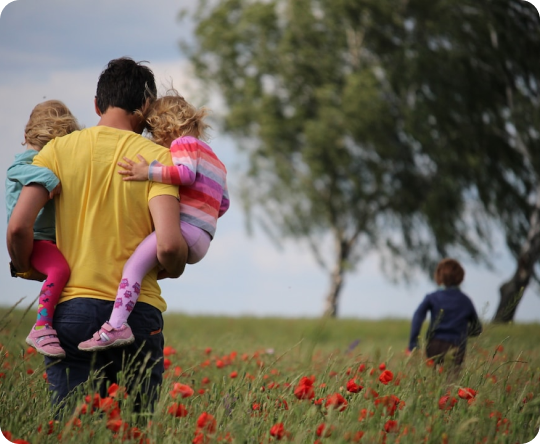 Backside of Man Holding Two Children In Each Arm With A Third Child In the Distance